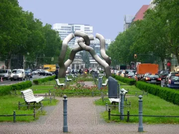 The Berlin sculpture of segmented grey tubes stands in a tree-lined street with cars, benches, and urban buildings nearby.