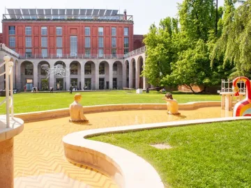 The Mystery Baths fountain with sculpted figures and a colorful swan in front of a red brick building and trees.