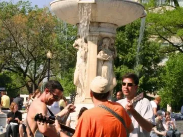 Dupont Circle Fountain with water flowing from the upper bowl and people gathered around it, including musicians in the foreground.