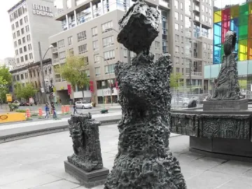 Dark bronze sculptures from La Joute in an urban plaza, with a large abstract figure in the foreground and a fountain base behind it.