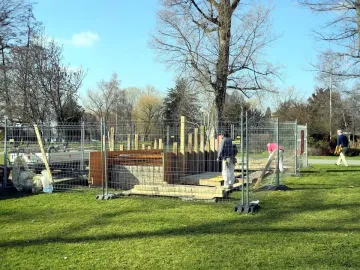 An outdoor construction site in a grassy park, fenced off with temporary metal barriers, with a partly built wooden and rebar structure and a few people nearby.