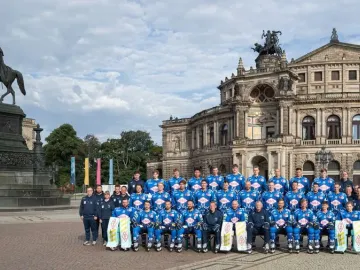 King Johann Memorial standing on a high stone pedestal in a public square, with ice hockey players posing near the opera house in the background.