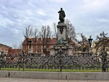 Adam Mickiewicz Monument showing a bronze cloaked figure on an ornate stone pedestal, with bare trees and wrought-iron fencing nearby.