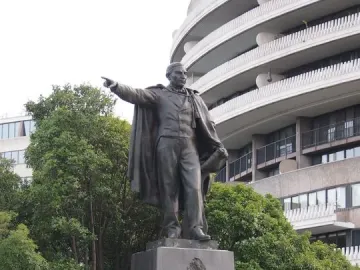 Bronze statue of Benito Juarez on a dark pedestal, pointing forward with a modern building and trees behind it.