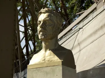 Gustave Eiffel bust on a pedestal with the Eiffel Tower structure in the background.