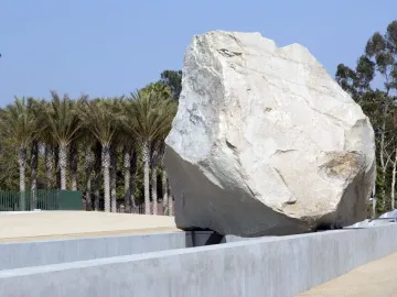 Levitated Mass sculpture with a large boulder appearing to hover above a concrete trough, with palm trees and blue sky behind it.
