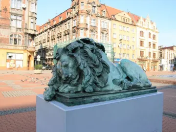 Sleeping Lion bronze statue with a green patina on a gray pedestal in a town square.