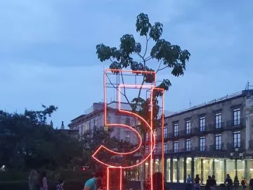 Antimonumento 5J glowing orange in a public plaza at twilight, with people and audio equipment nearby.