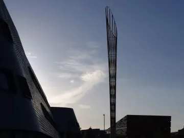 Aspire sculpture silhouetted against a bright, partly cloudy sky near modern campus buildings.