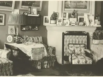 A small dark crouching Venus statuette sits on a crowded mantelpiece beside a clock and framed photographs.