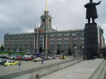 Lenin Monument seen from behind with one arm raised, in front of a large historic building and parked cars.