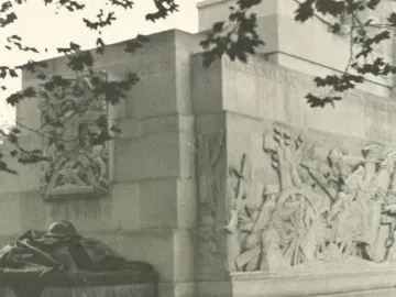 Royal Artillery Memorial with stone relief carvings and a helmet sculpture on a lower platform, partly framed by tree branches.