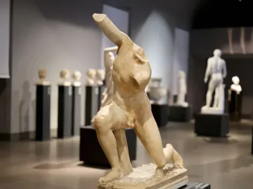Damaged marble sculpture of the Subiaco Ephebe with missing head, arms, and lower legs, displayed on a pedestal in a museum hall.