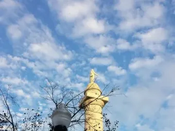 The Washington Monument stone column rises above trees and a streetlamp against a bright blue sky with scattered clouds.