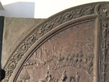 Ornate bronze gate panel with relief scenes of horsemen, figures in a landscape, a Latin inscription, and a crowded battle below.