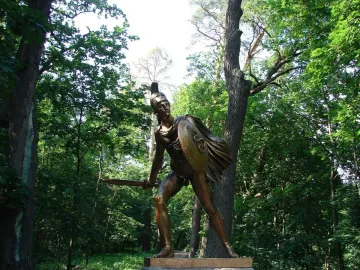 Greek Warrior Monument: a bronze armored warrior with a crested helmet, shield, and blade among tall trees.