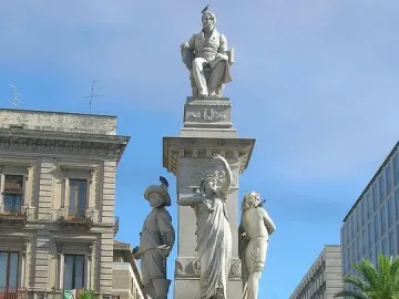 The Monument to Vincenzo Bellini, a tall white marble sculpture with a seated figure at the top and three standing allegorical figures below, surrounded by a decorative fence in a city square.