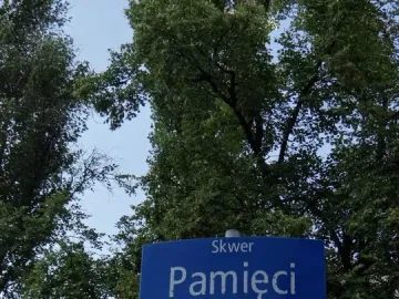 Blue Skwer Pamięci street sign mounted on a light-colored pole with trees and a building in the background.