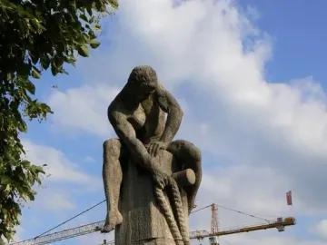 Tauzieher stone sculpture of a crouching figure fastening a thick rope to a post, with cranes and a blue sky behind it.