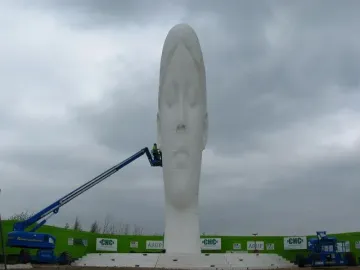 The Dream sculpture stands tall with a serene face under a cloudy sky, while a worker in a blue hydraulic lift works near the base.