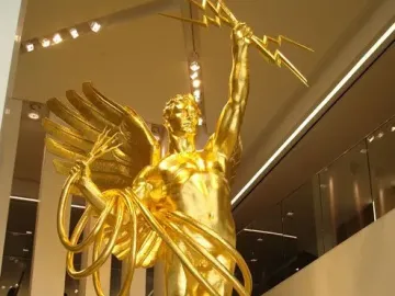 Golden Boy sculpture: a reflective golden winged man holding a lightning bolt and cable forms above a sphere