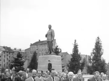A tall statue of Joseph Stalin stands on a multi-tiered pedestal in a public square, with several men walking in the foreground.