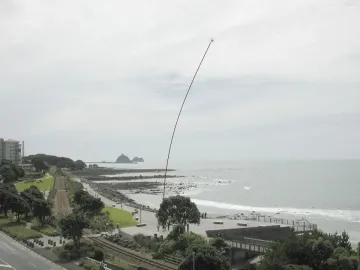 The Wind Wand sculpture rises from a coastal promenade beside the shoreline and city buildings under a blue sky.