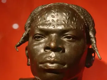 Dark bronze bust of an Aruwimi native with detailed hair or head markings against a red background.