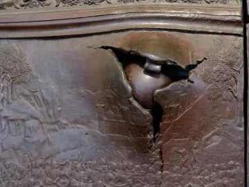 Close-up of the Bronze Gate in Naples, showing a jagged rupture with a rounded object lodged in the bronze panel.