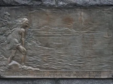 Bronze relief plaque on the Chief Seattle statue showing a figure looking toward the water, mountains, a rising sun, and a sailing ship.