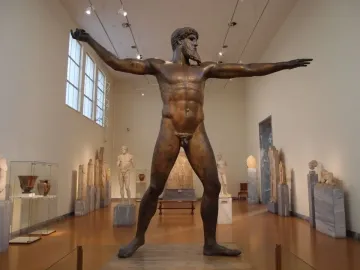The Artemision Bronze stands on display in a museum gallery, showing a bearded nude figure with both arms outstretched.
