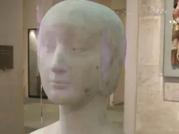 White marble bust of a princess with a serene expression and close-fitting cap on a dark base in a museum display.