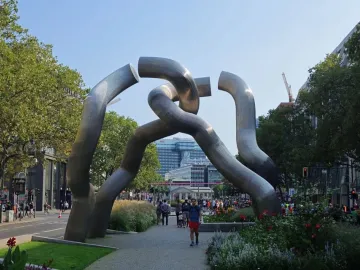 Intertwined shiny metal tube sculpture in a Berlin plaza with people and buildings nearby.