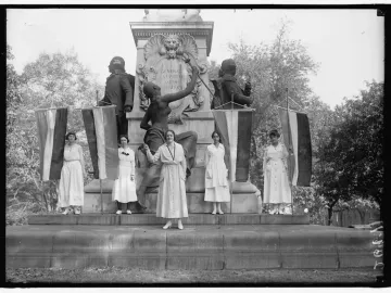 Women standing on the base of the Marquis de Lafayette Statue with suffrage flags in front of bronze figures and busts.