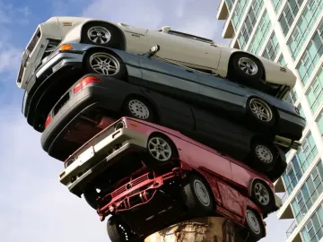 Trans Am Rapture sculpture with stacked cars rising from a large tree trunk against a blue sky, with a modern building in the background.