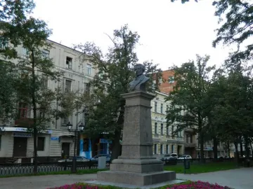 Bust of Pushkin atop a tall stone monument beside a flowerbed of pink blooms, with city buildings and trees behind it.