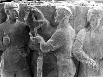 Weathered stone relief with three male figures in caps and uniforms around a central lightning-bolt symbol.