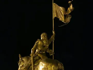 Golden Joan of Arc equestrian statue seen from behind-left, with a flowing banner and bright light beneath the horse.