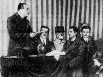 A man speaks to seated men beside a draped table in front of a striped backdrop.