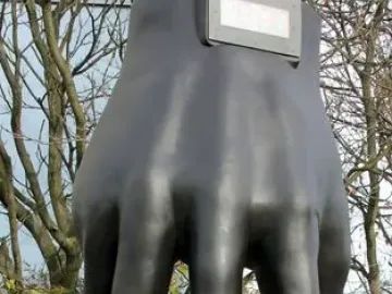 Hand with Clock sculpture on a tall pedestal beside a digital time display, with trees, bicycles, and a nearby building in the background.