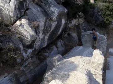 The unfinished Kouros of Apollonas лежing in a quarry, with a person standing beside its massive stone form.