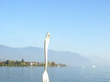 Fork of Vevey sculpture standing in the water of Lake Geneva near a rocky shore, with mountains and buildings in the distance.