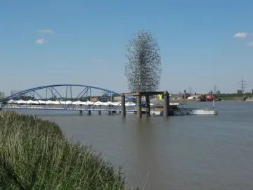 Quantum Cloud sculpture on a riverside platform beside a blue pedestrian bridge.