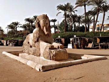 The Sphinx of Memphis on a low stone base in a sandy open area with palm trees and small stalls in the background.