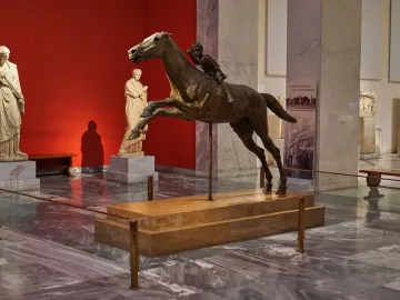 The Jockey of Artemision bronze sculpture shown with a horse and young rider on a pedestal in a museum gallery.