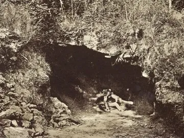 Sepia photograph of the cave entrance at Brassempouy, with a man seated in the dark opening surrounded by vegetation.