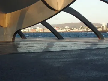 The Sun Voyager sculpture seen through its curved metallic forms, with blue water, a distant cityscape, and hills in the background.