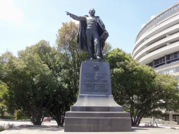 Bronze statue of Benito Juarez standing on a tall inscribed pedestal with trees and a curved building in the background.