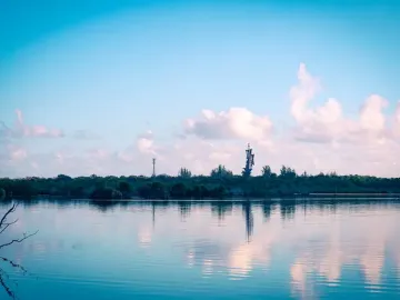 Birth of the New World sculpture reflected in calm water near a tree-lined shore.