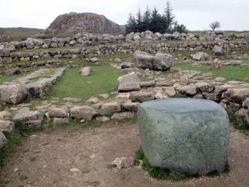 Large greenish stone block in the foreground at Hattusa, with ruins of stone walls and a rocky hill behind it.